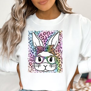 May include: White sweatshirt with a bunny face graphic wearing glasses and a bow. The bunny is set against a colourful leopard print background. The bunny's face is white with black outlines, and the glasses are black.