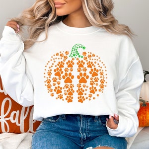 May include: White sweatshirt featuring an orange pumpkin design made of paw prints. The pumpkin stem is green. The sweatshirt is part of a fall-themed collection. An orange pillow with the words "happy fall" is visible.
