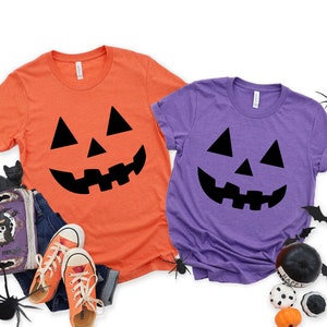 May include: Two t-shirts with a black jack-o'-lantern face design. One shirt is orange and the other is purple.