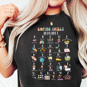 May include: A black t-shirt with a white graphic of a colorful alphabet chart. The chart lists coping skills for each letter of the alphabet. The text reads "Coping Skills Alphabet".