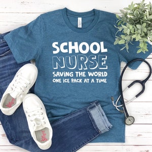 May include: A teal blue t-shirt with the text "SCHOOL NURSE SAVING THE WORLD ONE ICE PACK AT A TIME" in white. A stethoscope is lying on a white wooden surface next to the shirt.