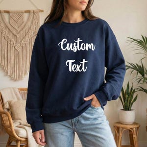 A woman wearing a navy blue sweatshirt with the text "Custom Text" printed on it, standing in a room with a woven wall hanging and potted plants in the background.