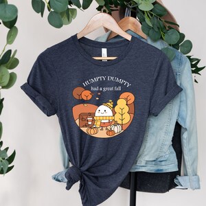 May include: A dark heather grey t-shirt with a cartoon illustration of a smiling egg wearing a hat and scarf. The text "Humpty Dumpty had a great fall" is printed on the shirt.