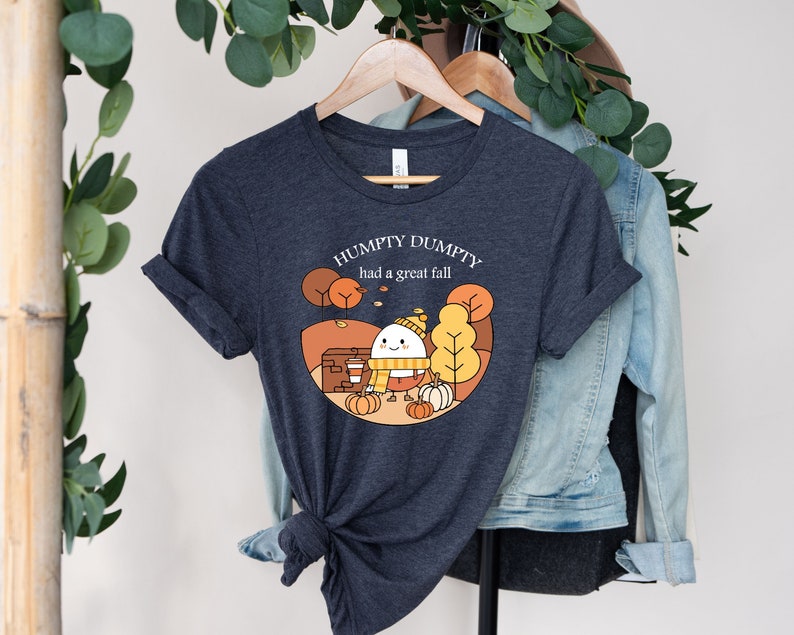May include: A dark heather grey t-shirt with a cartoon illustration of a smiling egg wearing a hat and scarf. The text "Humpty Dumpty had a great fall" is printed on the shirt.