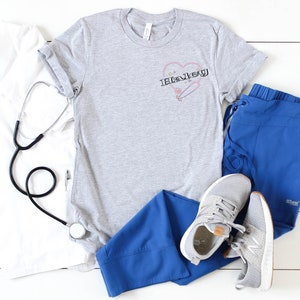 K&ouml;nnte beinhalten: Ein graues T-Shirt mit einem Herzdesign, das das Wort "Haley" und eine Sicherheitsnadel zeigt. Das Shirt liegt auf einer wei&szlig;en Oberfl&auml;che mit einem Stethoskop, einem wei&szlig;en Kittel, einer blauen Scrubbhose und einem Paar grauen Sneakers.