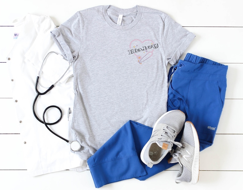 K&ouml;nnte beinhalten: Ein graues T-Shirt mit einem Herzdesign, das das Wort "Haley" und eine Sicherheitsnadel zeigt. Das Shirt liegt auf einer wei&szlig;en Oberfl&auml;che mit einem Stethoskop, einem wei&szlig;en Kittel, einer blauen Scrubbhose und einem Paar grauen Sneakers.