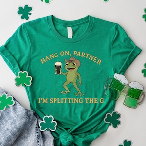 A green t-shirt with a cartoon frog holding a mug and the text "HANG ON PARTNER I'M SPLITTING THE G" printed on it, along with a pair of jeans and shamrocks.