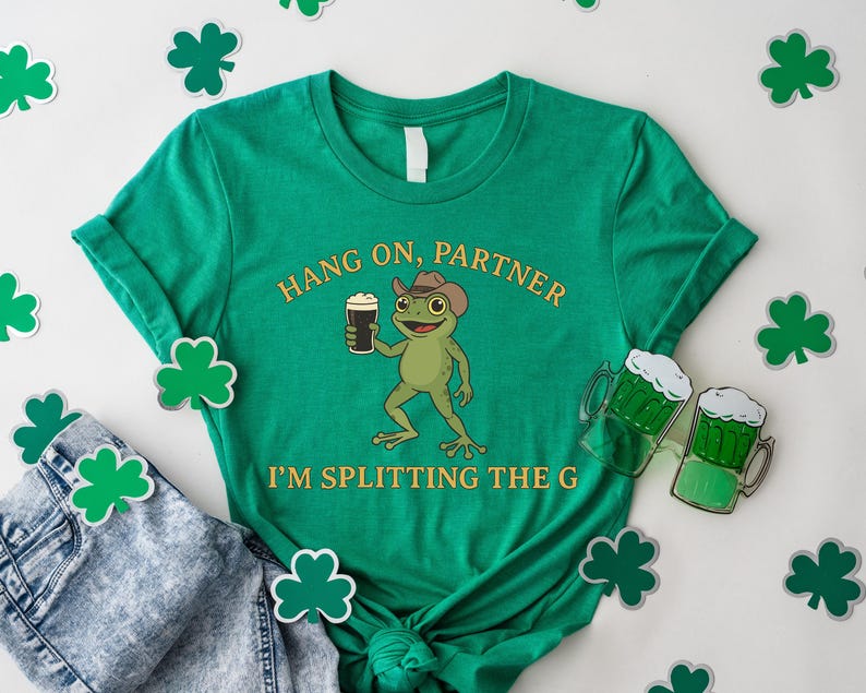 A green t-shirt with a cartoon frog holding a mug and the text "HANG ON PARTNER I'M SPLITTING THE G" printed on it, along with a pair of jeans and shamrocks.