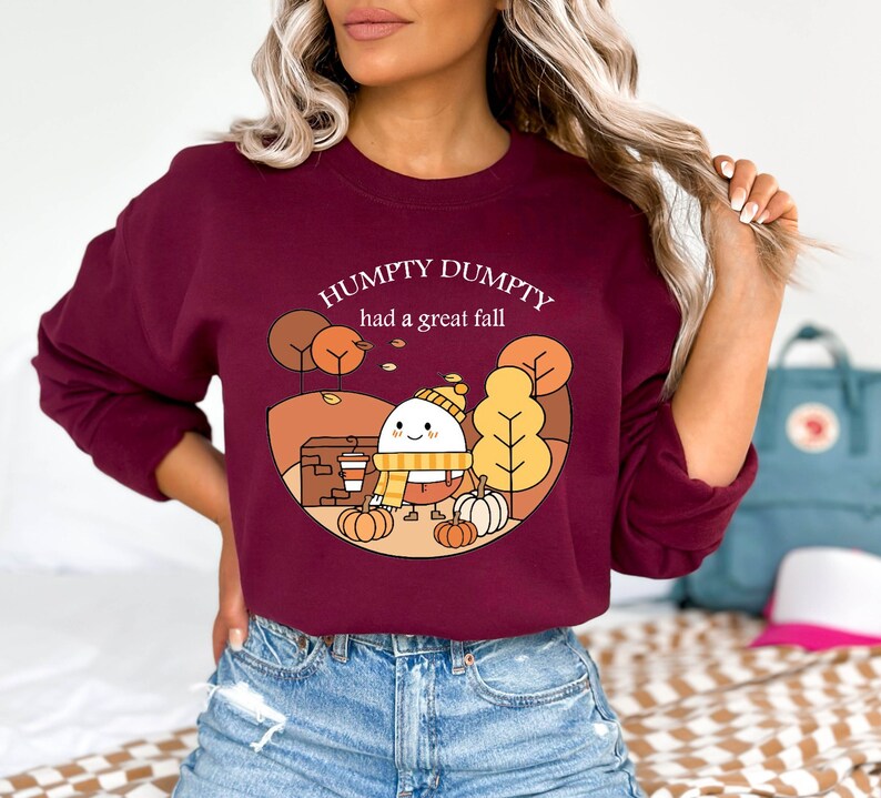 May include: A maroon long-sleeved sweatshirt with a cartoon illustration of an egg wearing a hat and scarf, holding a cup of coffee. The text "Humpty Dumpty had a great fall" is printed on the sweatshirt.