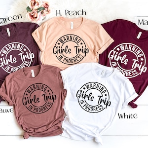 May include: Five t-shirts in different colors with the text "WARNING Girls Trip In Progress" in a circle on the front. The colors of the shirts are maroon, peach, white, mauve, and maroon.