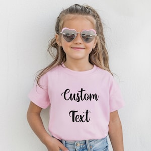A young girl wearing a pink t-shirt with the text "Custom Text" printed on it, smiling and wearing heart-shaped sunglasses.