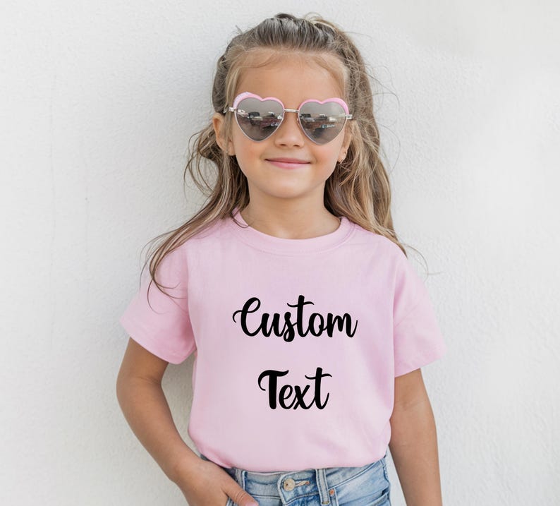 A young girl wearing a pink t-shirt with the text "Custom Text" printed on it, smiling and wearing heart-shaped sunglasses.