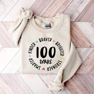 Op de afbeelding: Een beige sweatshirt met een zwarte cirkelvormige afbeelding waarop "100 dagen" staat en die is omringd door de woorden "Kinder", "Smarter", "Stronger", "Brighter" en "Braver" met kleurrijke hartjes.