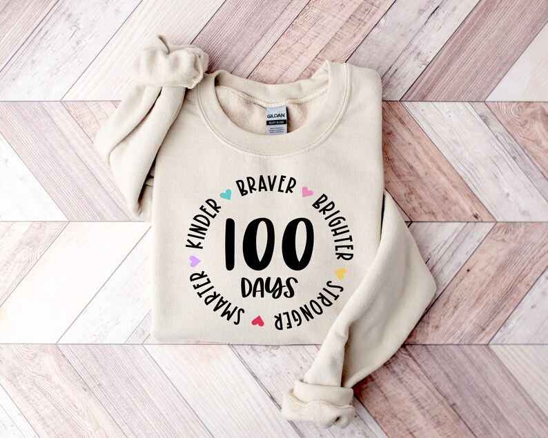 Op de afbeelding: Een beige sweatshirt met een zwarte cirkelvormige afbeelding waarop "100 dagen" staat en die is omringd door de woorden "Kinder", "Smarter", "Stronger", "Brighter" en "Braver" met kleurrijke hartjes.