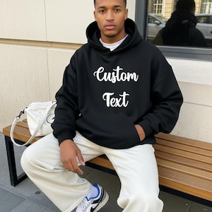 A man wearing a black hoodie with white text sits on a bench.