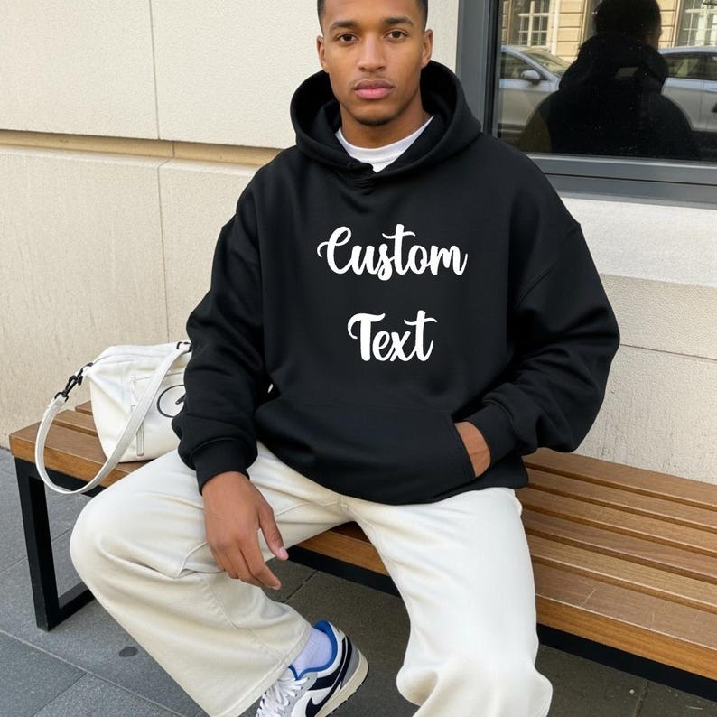 A man wearing a black hoodie with white text sits on a bench.