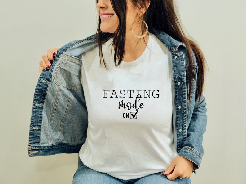 May include: White t-shirt with the text "FASTING mode on" in black script. The shirt is being worn under a denim jacket. The text includes a checkmark symbol.