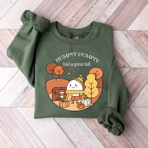 May include: Green sweatshirt with a cartoon illustration of a smiling egg wearing a hat and scarf, holding a cup of coffee and surrounded by pumpkins. The text "Humpty Dumpty had a great fall" is printed on the sweatshirt.