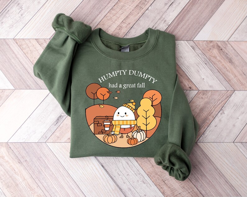 May include: Green sweatshirt with a cartoon illustration of a smiling egg wearing a hat and scarf, holding a cup of coffee and surrounded by pumpkins. The text "Humpty Dumpty had a great fall" is printed on the sweatshirt.