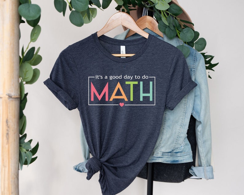 May include: A dark blue t-shirt with the text "it's a good day to do MATH" in a colorful rainbow font. The shirt is hanging on a hanger with a denim jacket.