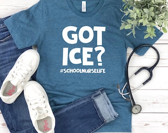 Peut inclure: Un t-shirt bleu sarcelle avec du texte blanc qui dit "GOT ICE? #SCHOOLNURSELIFE". Le t-shirt est posé à plat sur une surface en bois blanc avec une paire de baskets blanches et un jean bleu.