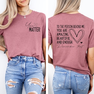 May include: A mauve t-shirt with the text "You Matter" on the front and the text "To the person behind me: You are amazing, beautiful and enough. Remember that." on the back. The shirt is being modeled by a person wearing blue jeans.