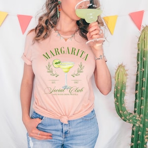 Puede incluir: Camiseta rosa claro con la inscripci&oacute;n "Margarita Social Club" y un dibujo de un vaso de margarita. La camiseta tiene las mangas dobladas y se combina con unos vaqueros azules. Una persona sostiene un vaso de margarita.