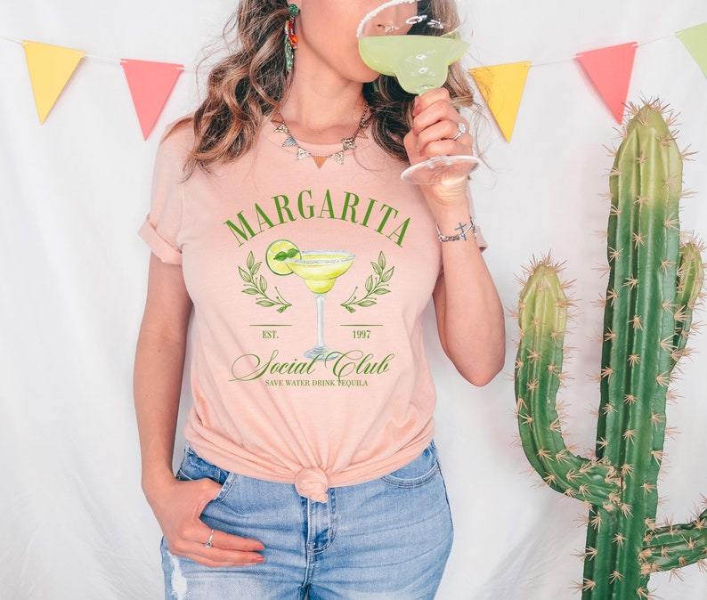Puede incluir: Camiseta rosa claro con la inscripci&oacute;n "Margarita Social Club" y un dibujo de un vaso de margarita. La camiseta tiene las mangas dobladas y se combina con unos vaqueros azules. Una persona sostiene un vaso de margarita.