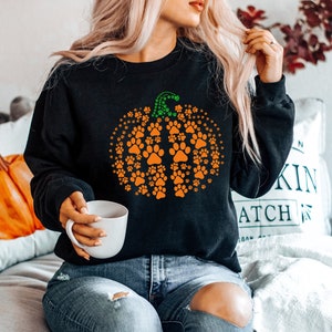 May include: Black sweatshirt featuring a pumpkin design made of orange paw prints with a green stem. The sweatshirt is a cozy fall-themed apparel item, perfect for pet lovers and autumn celebrations.