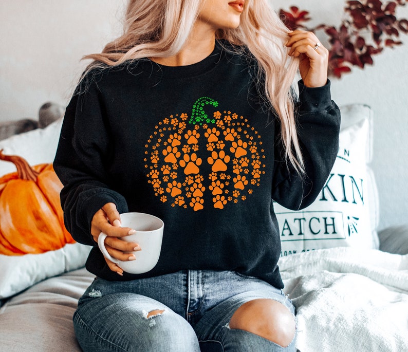 May include: Black sweatshirt featuring a pumpkin design made of orange paw prints with a green stem. The sweatshirt is a cozy fall-themed apparel item, perfect for pet lovers and autumn celebrations.