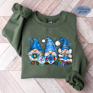 May include: A green sweatshirt with a Christmas gnome design. Three gnomes are wearing blue hats and holding a snowman, snowflake, and a cup of hot cocoa. The gnomes are surrounded by snowflakes.