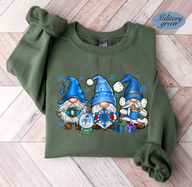 May include: A green sweatshirt with a Christmas gnome design. Three gnomes are wearing blue hats and holding a snowman, snowflake, and a cup of hot cocoa. The gnomes are surrounded by snowflakes.