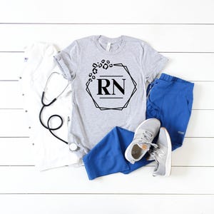May include: A gray t-shirt with a black geometric design and the letters "RN" is displayed with blue scrubs, gray sneakers, a white lab coat, and a stethoscope. The t-shirt design includes leopard print accents.
