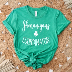 May include: A green t-shirt with the words "Shenanigans Coordinator" in white script and a white shamrock graphic. The shirt is knotted at the bottom and rests on a woven, circular rug.