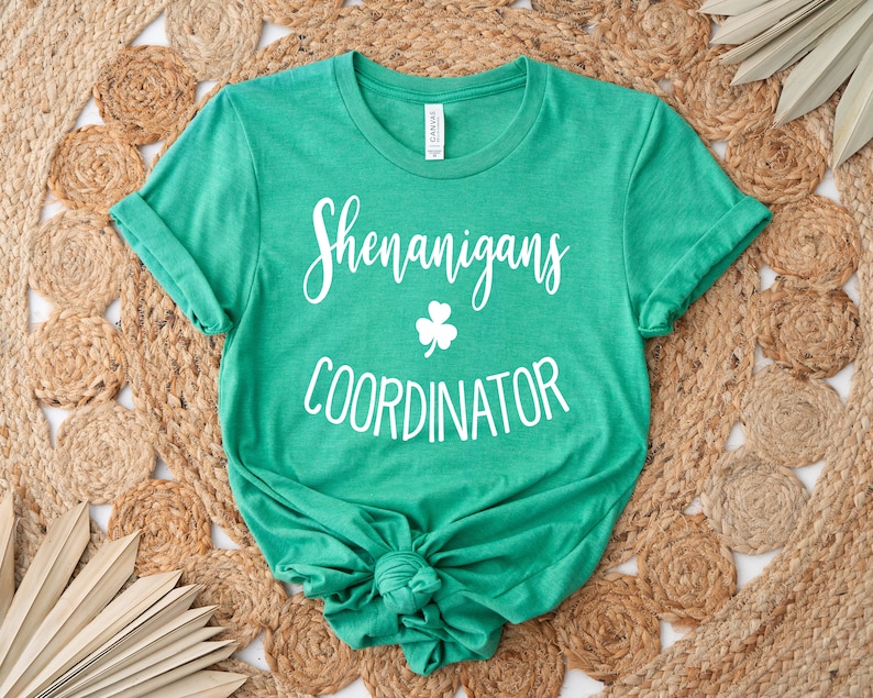 May include: A green t-shirt with the words "Shenanigans Coordinator" in white script and a white shamrock graphic. The shirt is knotted at the bottom and rests on a woven, circular rug.