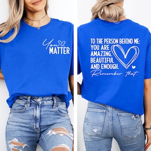 May include: A royal blue t-shirt with white text that reads "You Matter" on the front and "To the person behind me: You are amazing, beautiful and enough. Remember that." on the back. The shirt is being modeled by a person wearing blue jeans.