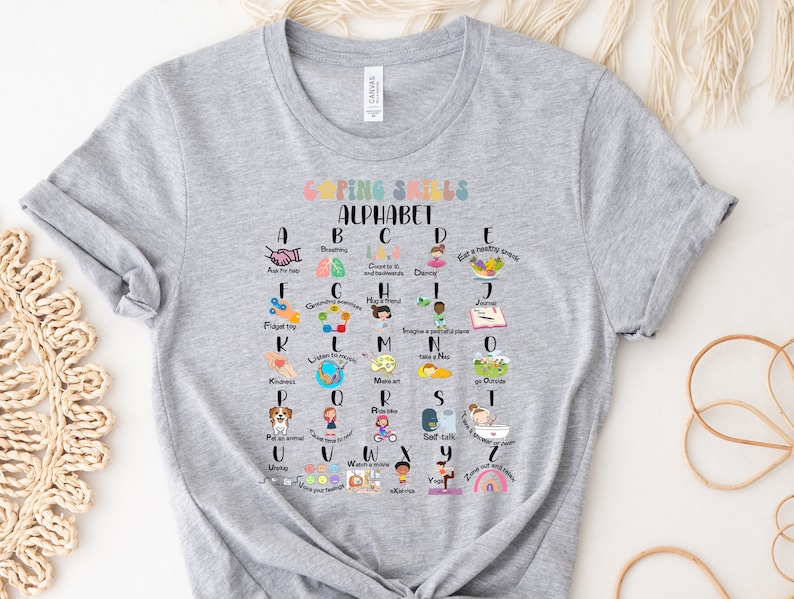 May include: A gray t-shirt with a colorful alphabet chart featuring coping skills for children. Each letter of the alphabet is paired with a coping skill, such as breathing, dancing, and taking a nap. The chart is surrounded by a white border.