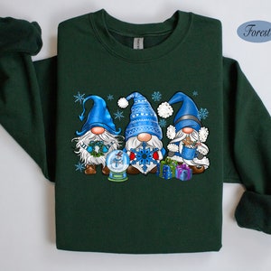 May include: A forest green sweatshirt with a Christmas gnome design. Three gnomes are wearing blue hats and holding gifts. The gnomes are standing in front of a snow globe with a snowflake.