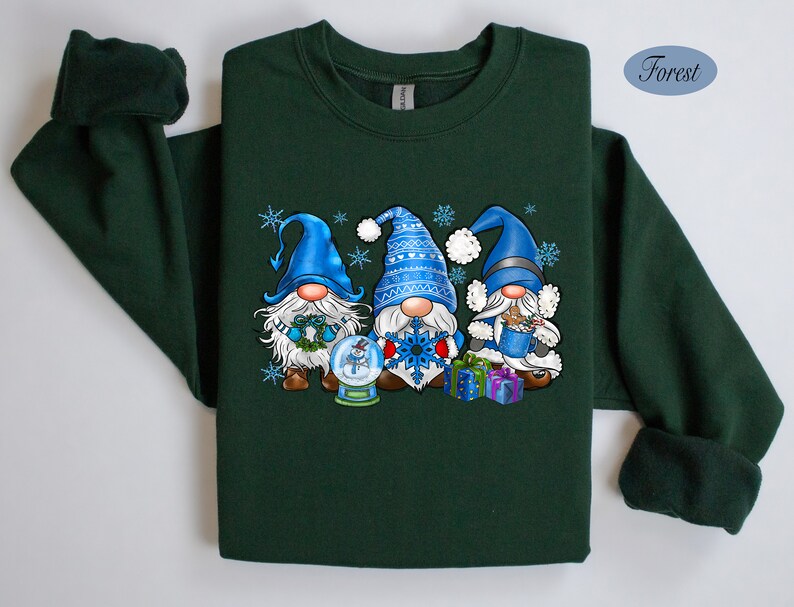 May include: A forest green sweatshirt with a Christmas gnome design. Three gnomes are wearing blue hats and holding gifts. The gnomes are standing in front of a snow globe with a snowflake.