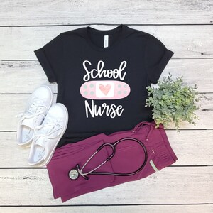 May include: Black t-shirt with the text "School Nurse" and a pink bandage with a heart on it. A stethoscope and scrubs are also pictured.