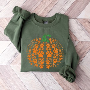 May include: An olive green sweatshirt featuring a pumpkin design made of orange paw prints. The pumpkin stem is green. The sweatshirt is folded on a light wood grain surface.