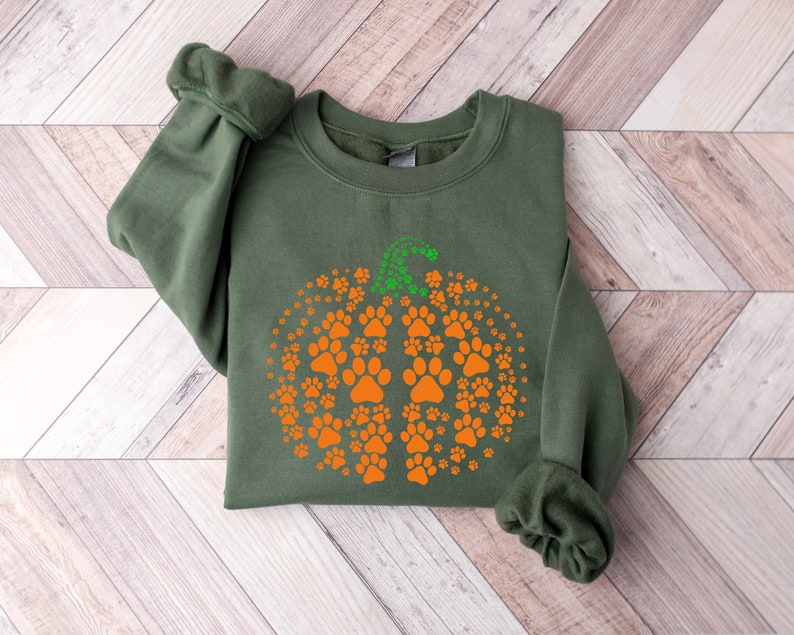 May include: An olive green sweatshirt featuring a pumpkin design made of orange paw prints. The pumpkin stem is green. The sweatshirt is folded on a light wood grain surface.