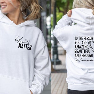 May include: White hoodie with a black graphic on the front that says "You Matter" with a heart symbol. The back of the hoodie has a black graphic that says "To the person behind me: You are amazing, beautiful and enough. Remember that." with a heart symbol.