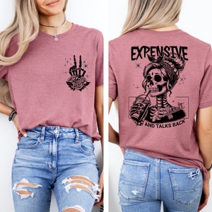 May include: A mauve t-shirt with a skeleton graphic on the back. The graphic shows a skeleton woman with a bandana, drinking from a cup. The text on the back reads "Expensive, Difficult, and Talks Back". The front of the shirt has a skeleton hand graphic with roses.