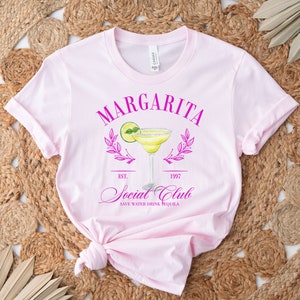 Puede incluir: Una camiseta rosa claro con un gr&aacute;fico de margarita. El gr&aacute;fico incluye la palabra "Margarita" encima de una ilustraci&oacute;n de c&oacute;ctel, con "Social Club" y "Save Water Drink Tequila" debajo. La camiseta est&aacute; sobre un fondo tejido.