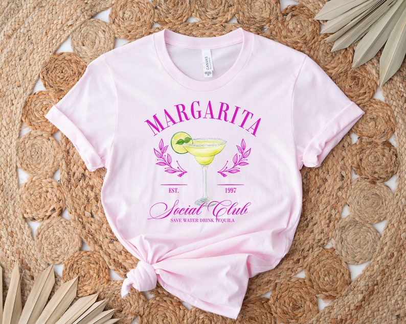 Puede incluir: Una camiseta rosa claro con un gr&aacute;fico de margarita. El gr&aacute;fico incluye la palabra "Margarita" encima de una ilustraci&oacute;n de c&oacute;ctel, con "Social Club" y "Save Water Drink Tequila" debajo. La camiseta est&aacute; sobre un fondo tejido.