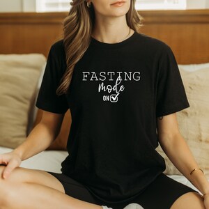 May include: Black t-shirt with the text "FASTING mode on" in white script and a checkmark. The shirt is short-sleeved and appears to be made of a soft material. The shirt is worn by a person sitting on a bed.