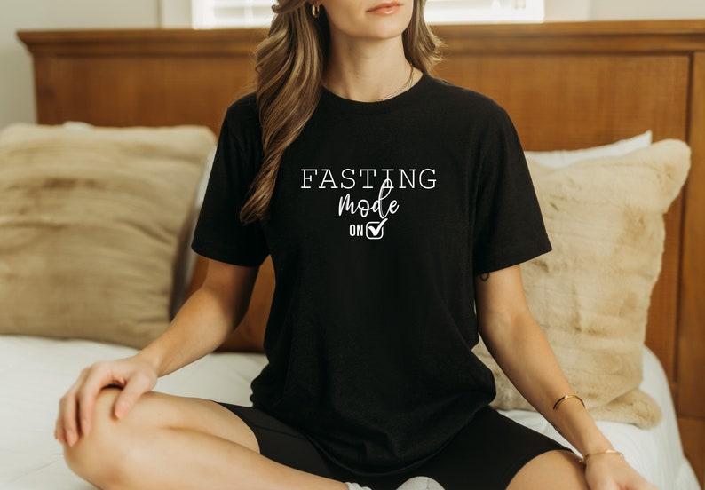 May include: Black t-shirt with the text "FASTING mode on" in white script and a checkmark. The shirt is short-sleeved and appears to be made of a soft material. The shirt is worn by a person sitting on a bed.