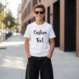 A young boy wearing a white t-shirt with the text "Custom Text" printed on it, standing on a sidewalk in front of a brick building.