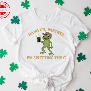 A white t-shirt with a cartoon frog holding a beer and the text "HANG ON PARTNER I'M SPLITTING THE G" printed on it. The t-shirt is surrounded by shamrocks on a white background.